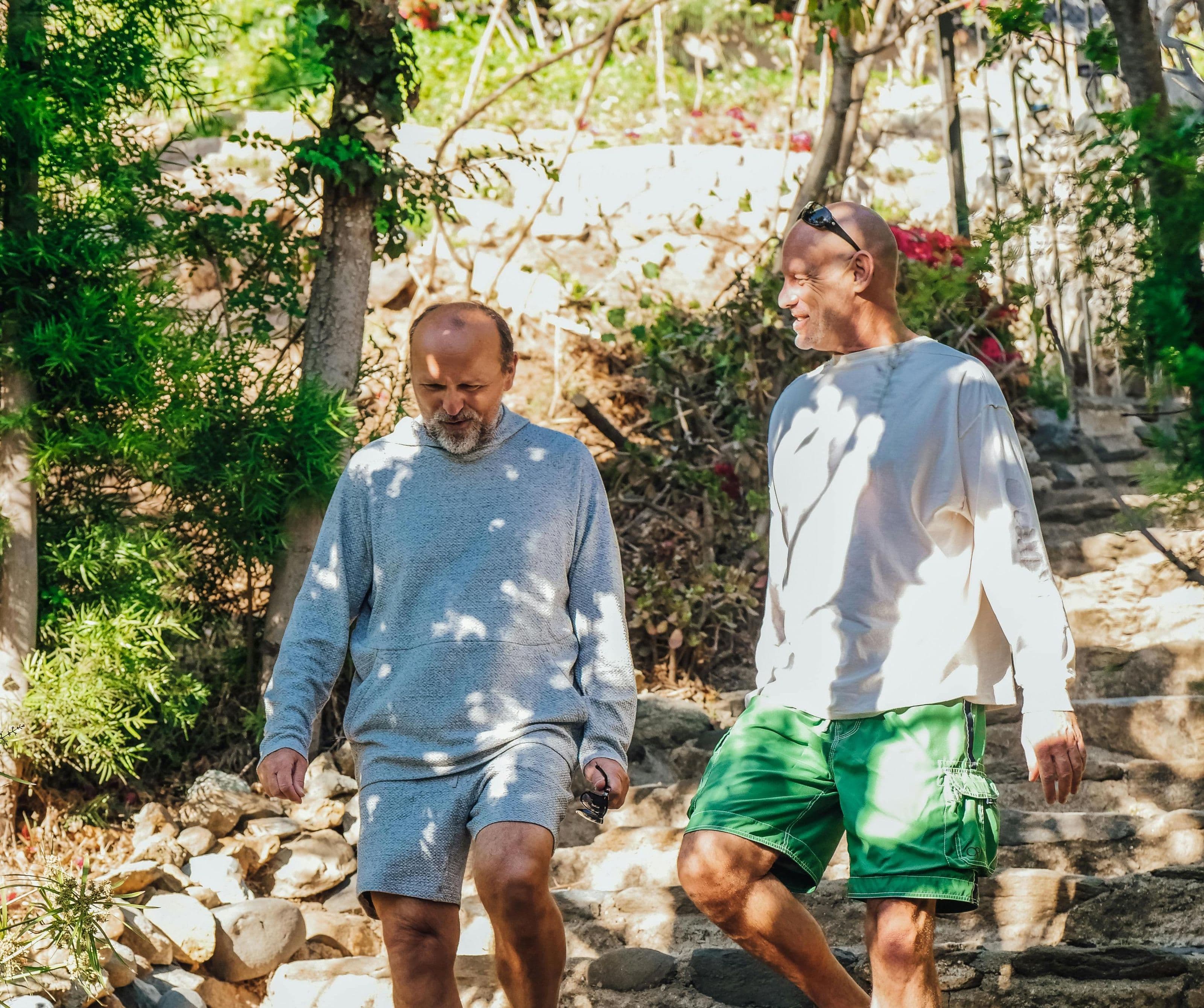 Two men in relaxed clothing walking and talking down sunlit stone garden steps surrounded by tropical greenery, reflecting the ongoing, supported integration work of the Afterlight AfterCare programme