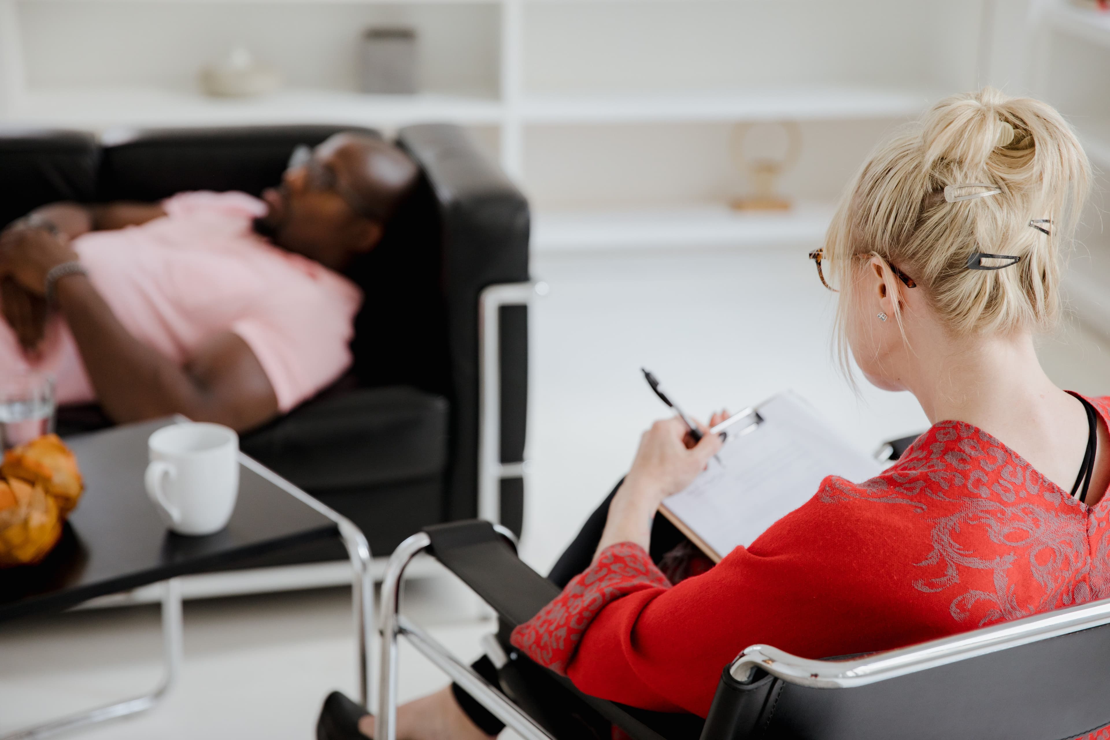 A therapist taking notes on a clipboard while a client reclines on a sofa during a clinical psychedelic therapy session