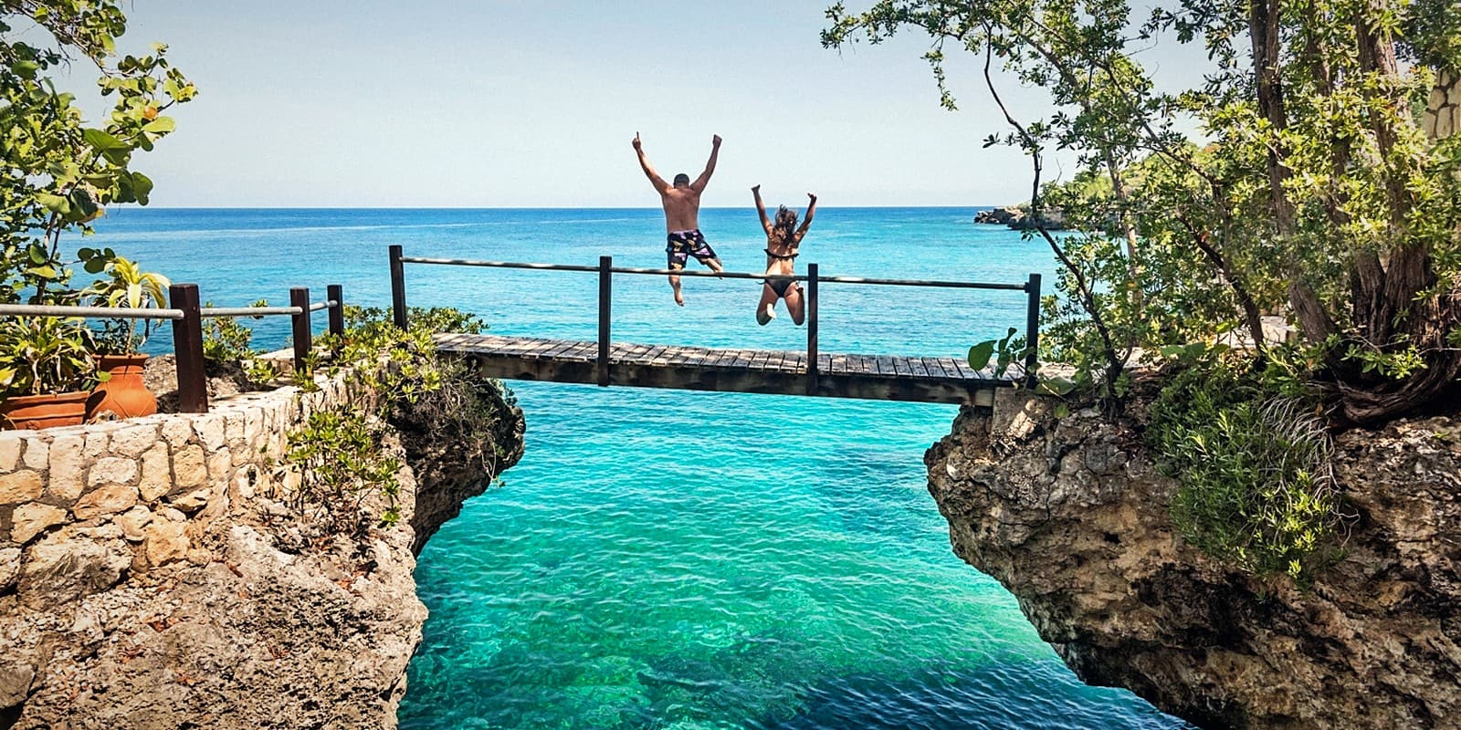 Two people leaping from a wooden footbridge into the turquoise water between the Negril cliffs