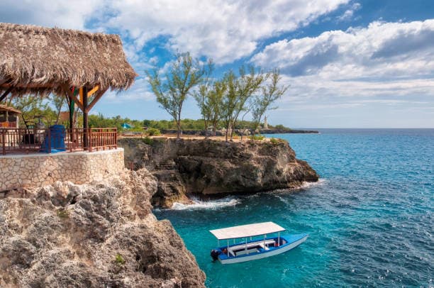 A thatched-roof hut on the limestone cliffs of Negril with a small boat moored in the turquoise cove below