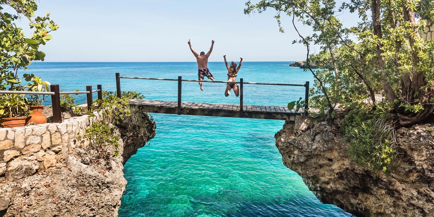 Two people leaping from a wooden footbridge into a turquoise cove between cliffs in Negril, Jamaica